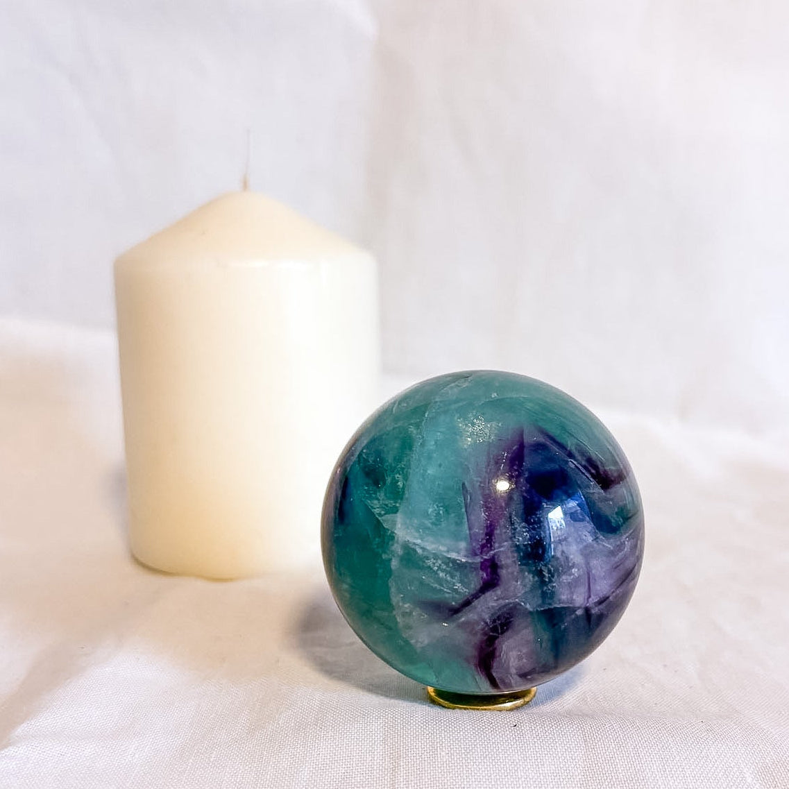 Fluorite green and purple crystal sphere next to a white candle on a white fabric background
