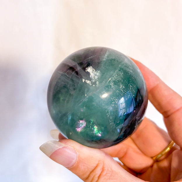 Hand holding Fluorite green and purple crystal sphere against a neutral background